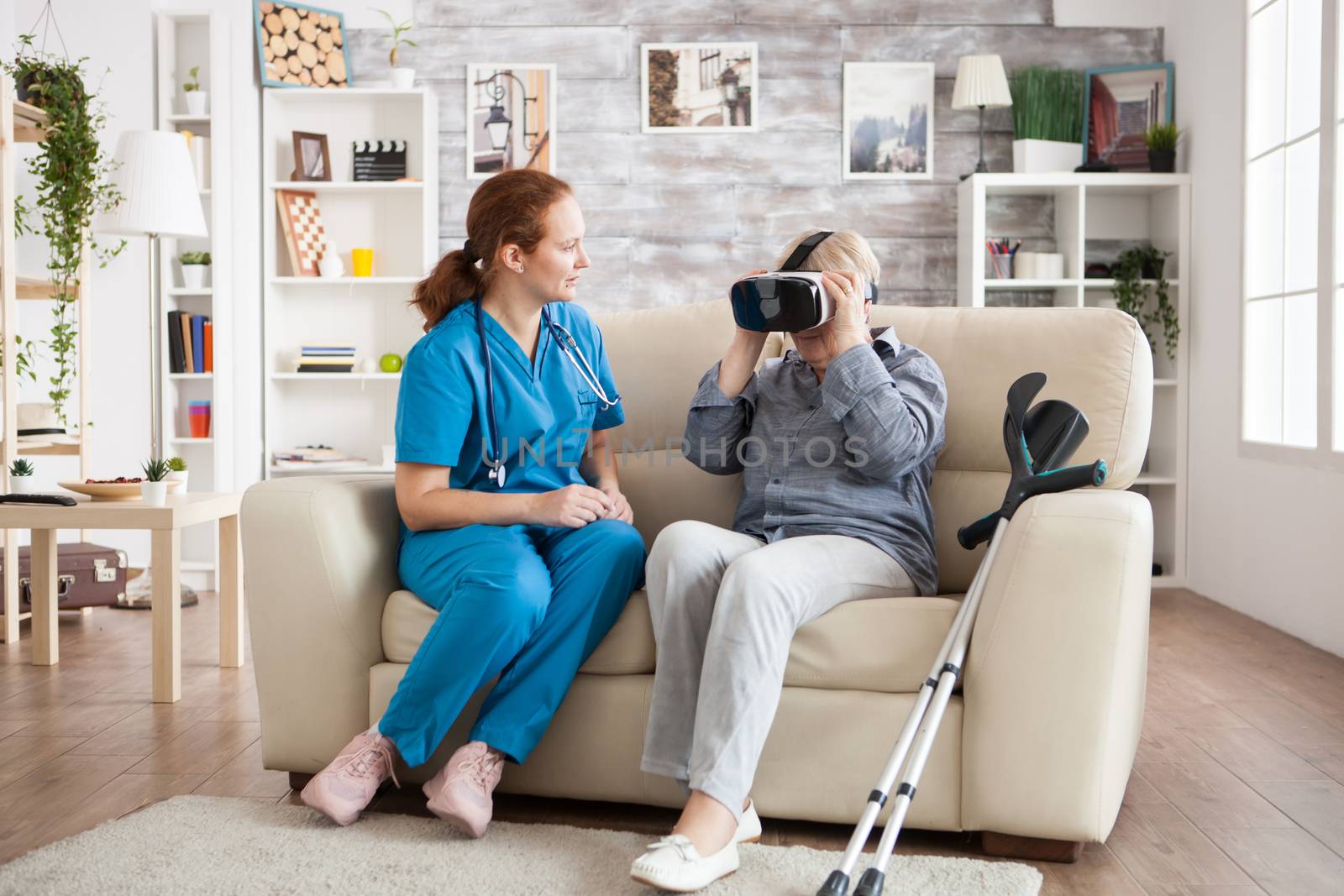 Old with with crutches in nursing home using virtual reality glasses with caregiver next to her.