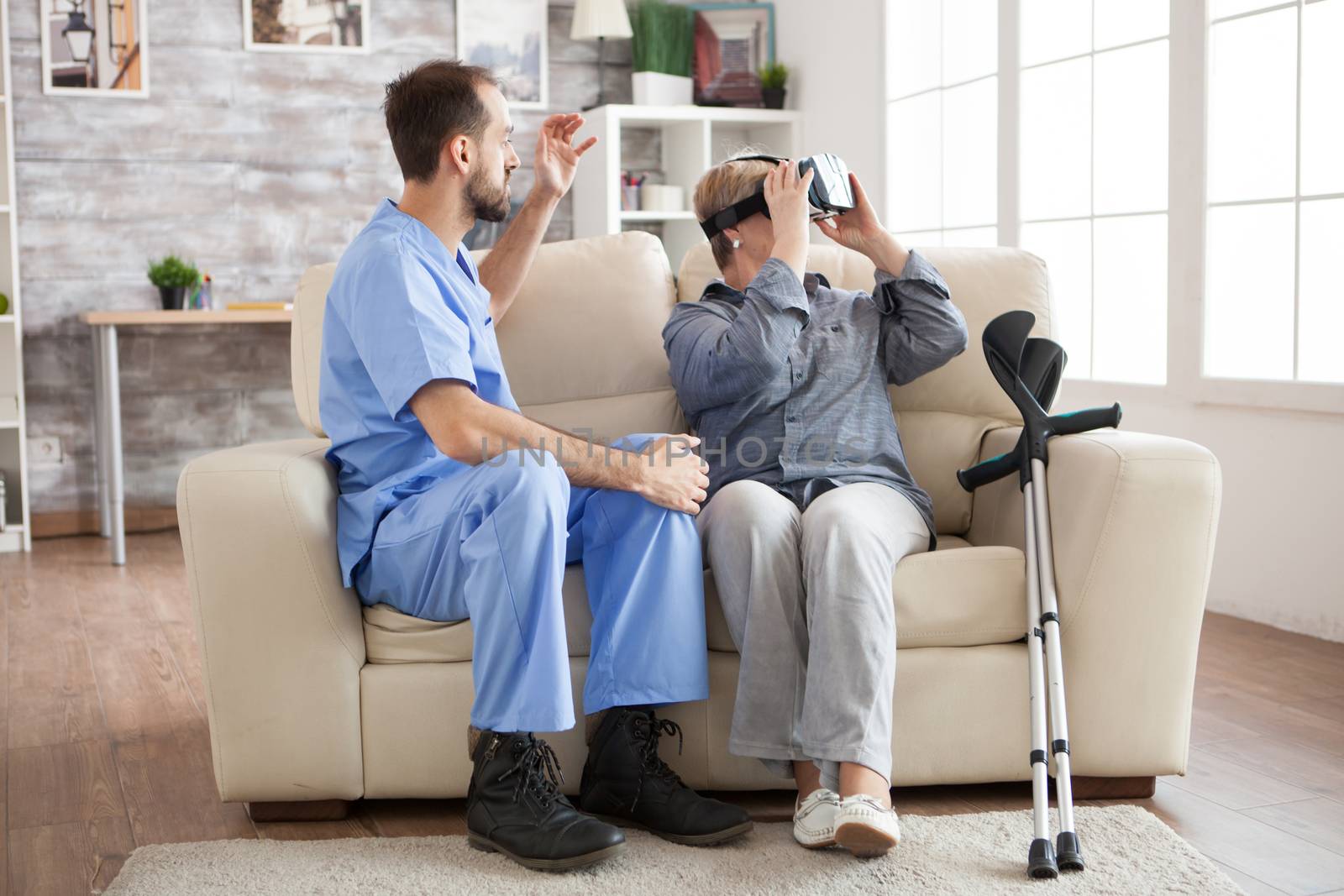 Caucasian old woman with crutches in nursing home using virtual reality glasses while doctor is talking about her diagnostic.