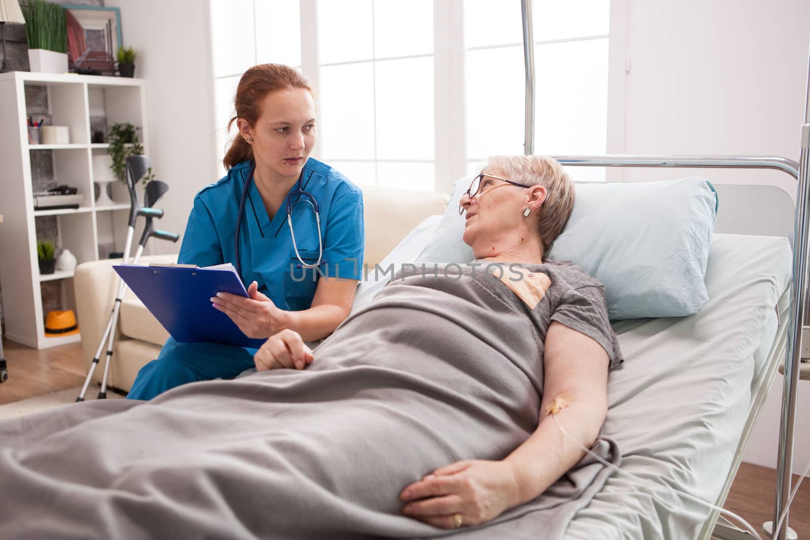 Old woman sitting in bed in nursing home talking with female doctor with clipboard.