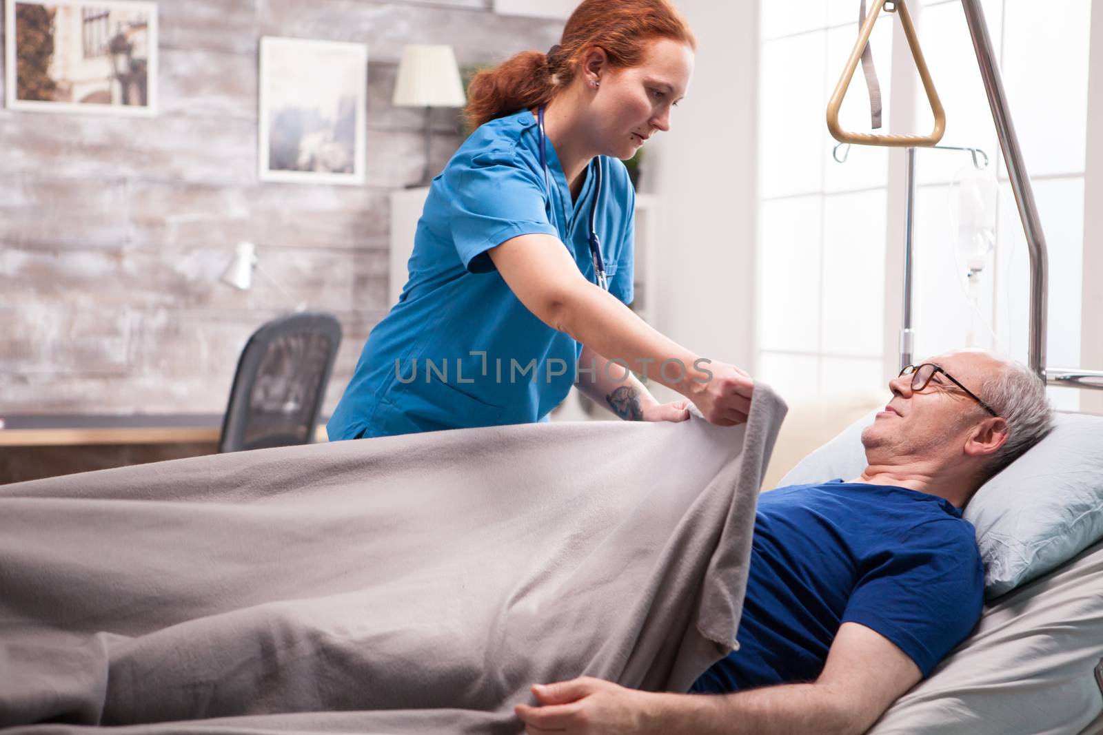 Female nurse tucking old man under the blanket in nursing home.