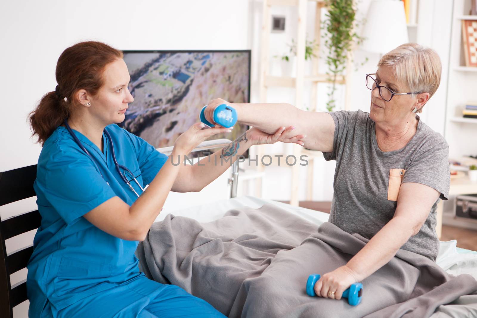 Caregiver helping using dumbbells for senior woman physiotherapy in nursing home.