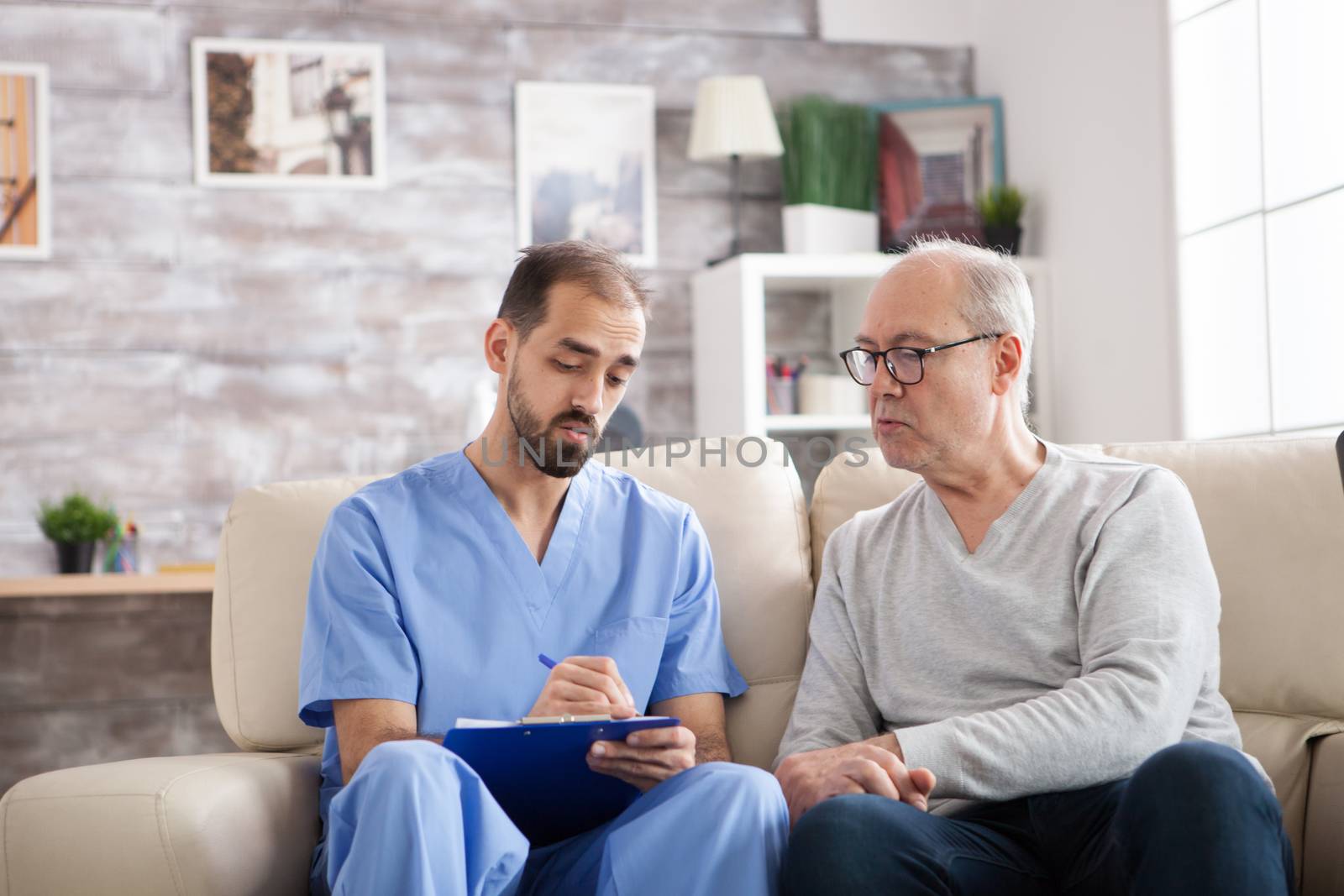 Male in his 60s sitting on couch in nursing home with doctor holding and writing on clipboard.