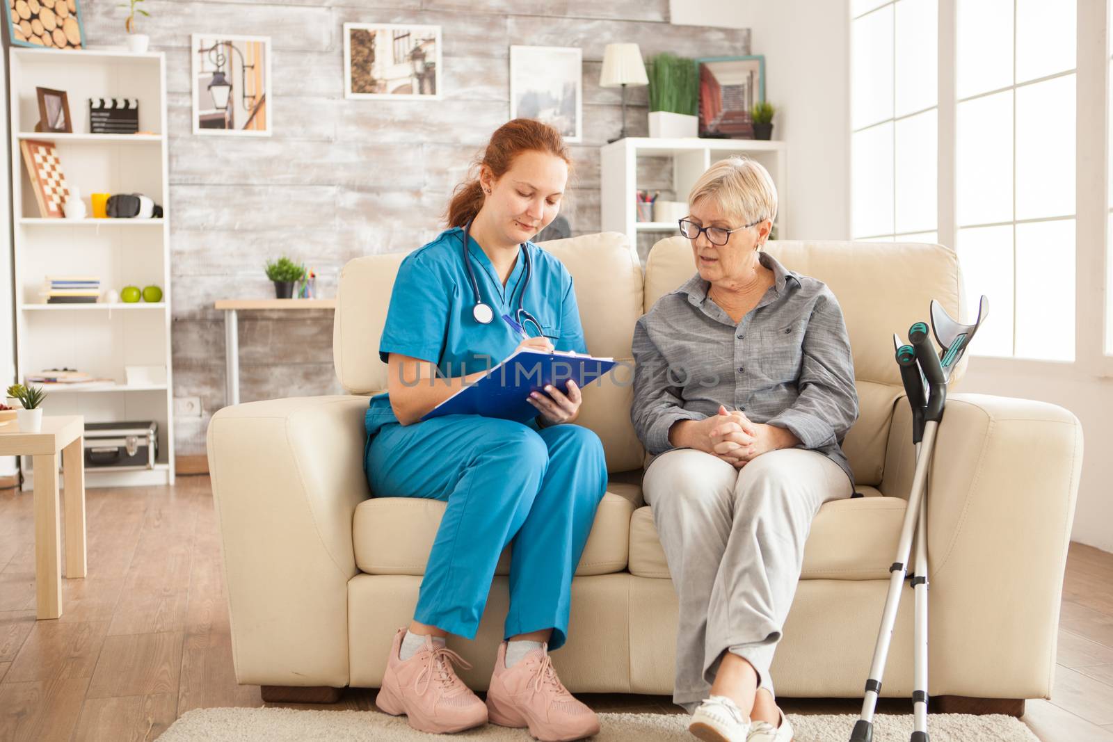 Female nurse writing on clipboard while talking with senior woman in nursing home.