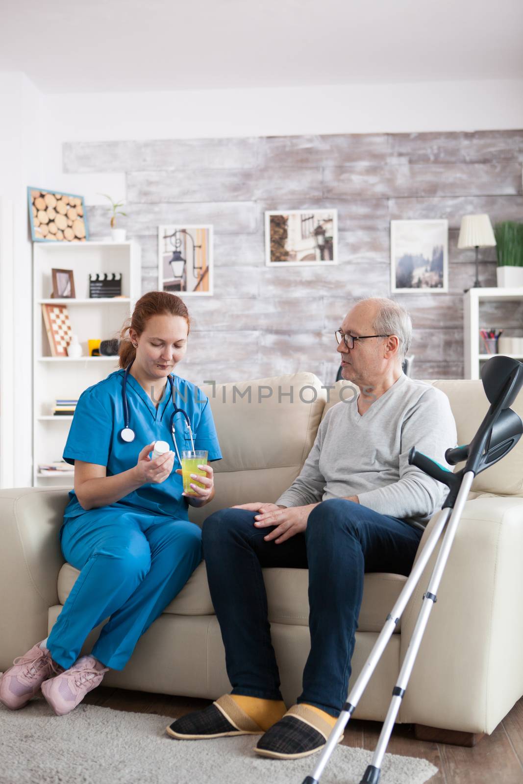 Pretty young nurse in nursing home giving medication to senior man with crutches.