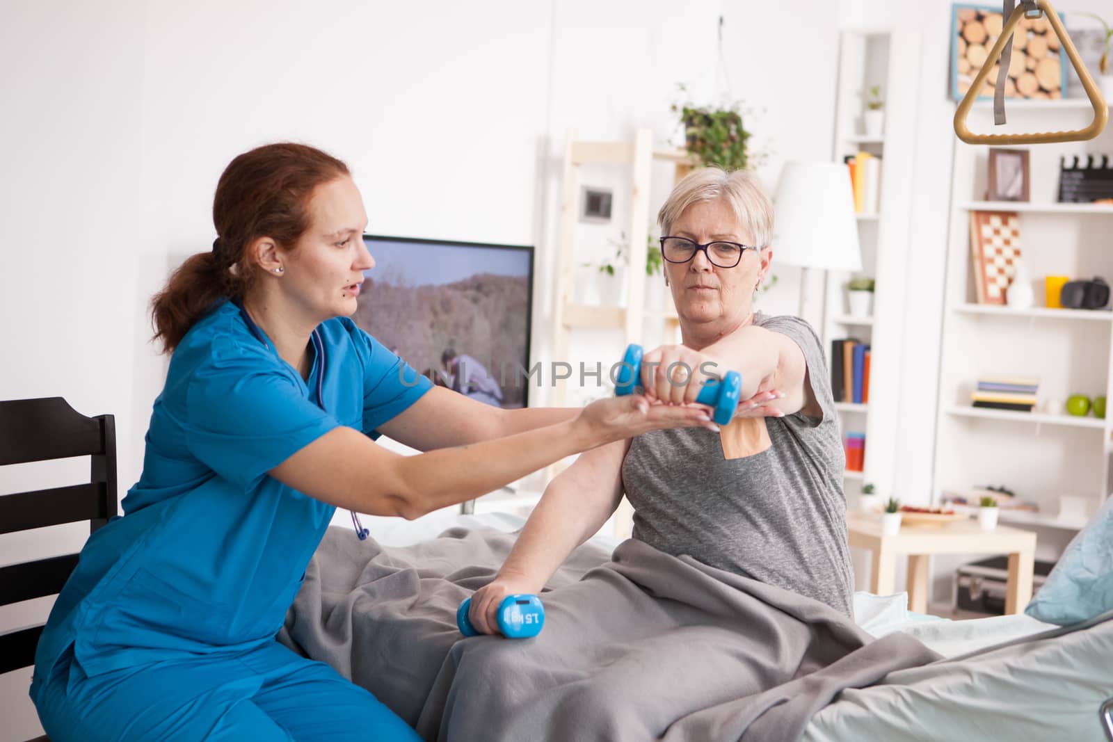 Female doctor helping elderly age woman with physiotherapy in nursing home with dumbbells.
