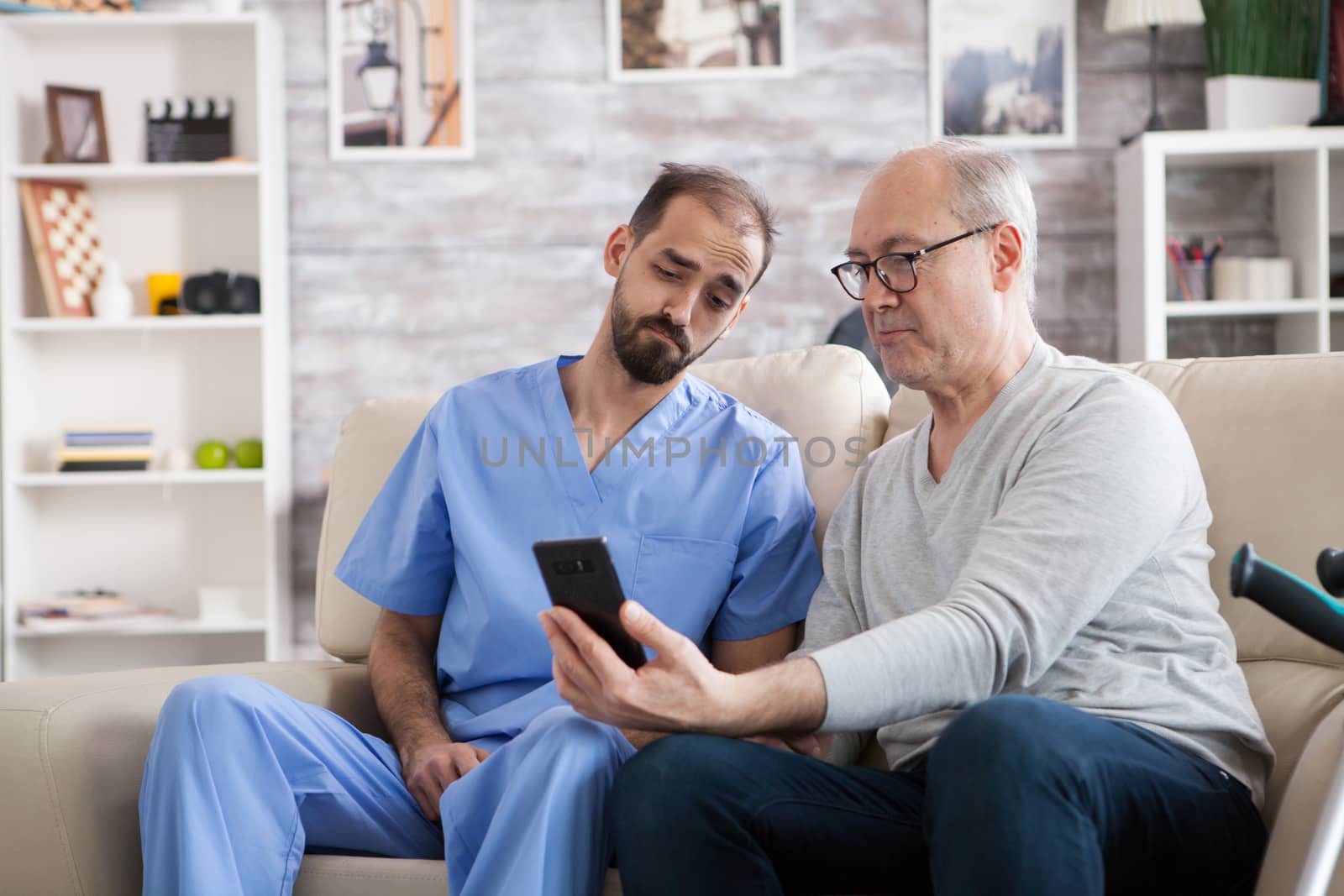 Retired man in modern nursing home sitting on couch with his health visitor. Senior man using mobile phone.