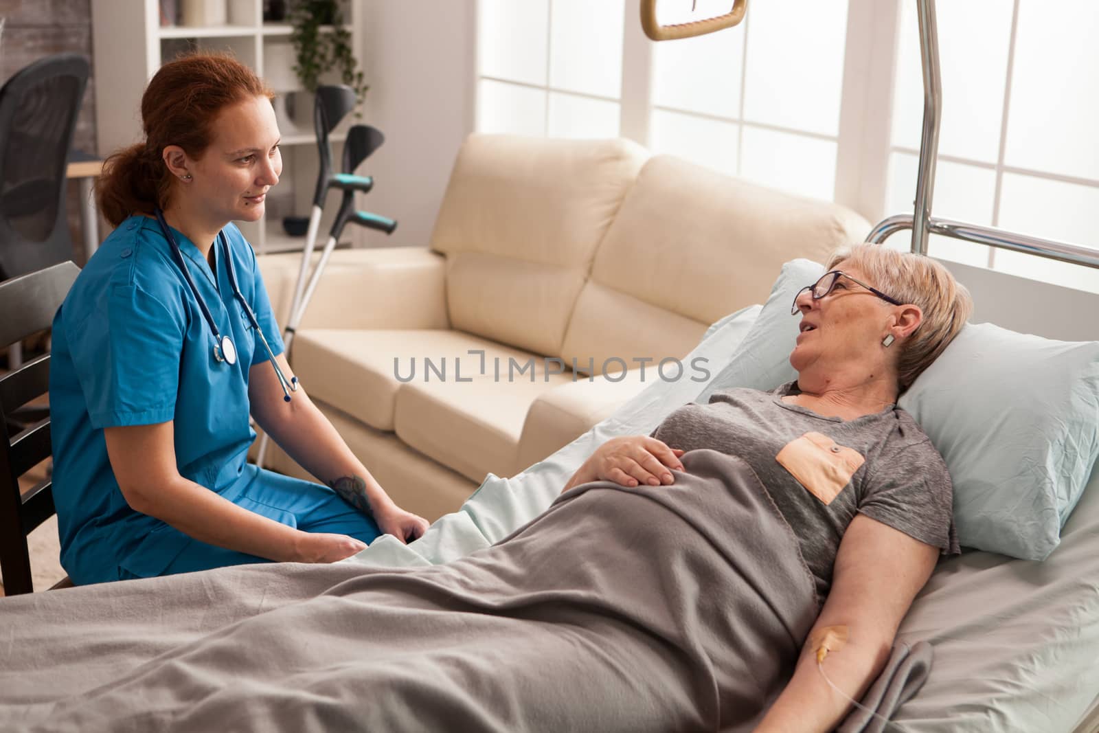 Ill senior woman laying on bed in a nursing home talking with female nurse.