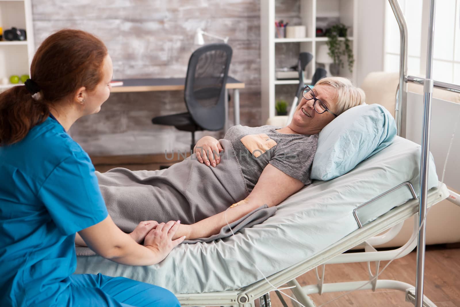 Female doctor in nursing home holding hand of old woman laying in bed.