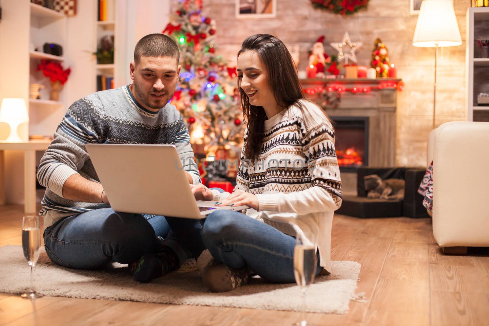 Relaxed couple on christmas day in front of fireplace smiling while using laptop.