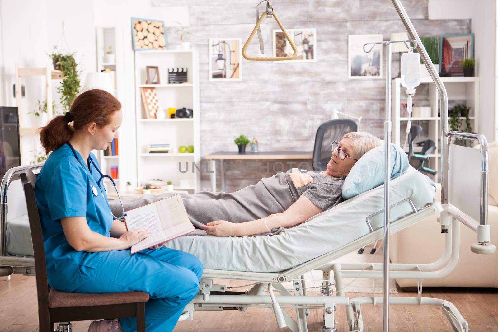 Female nursing sitting on chair next to old woman in nursing home reading a book.