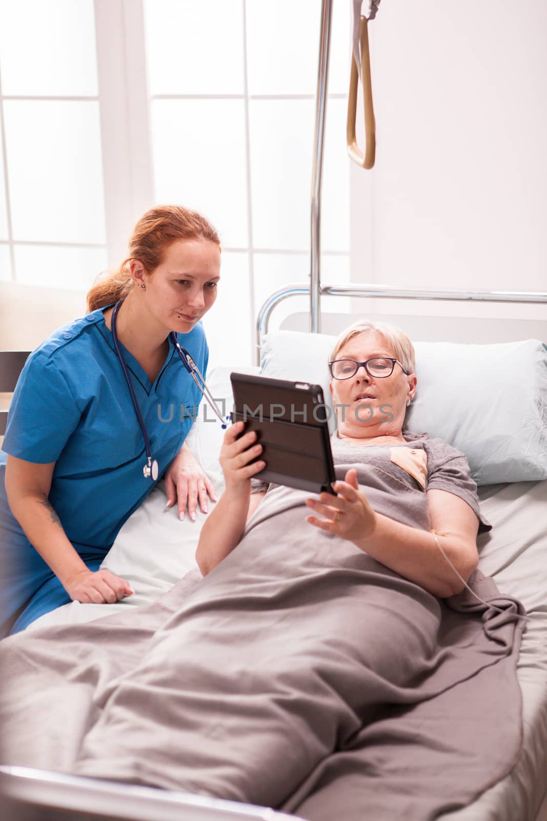 Senior woman lying in nursing home bed showing something to female nurse in tablet computer.