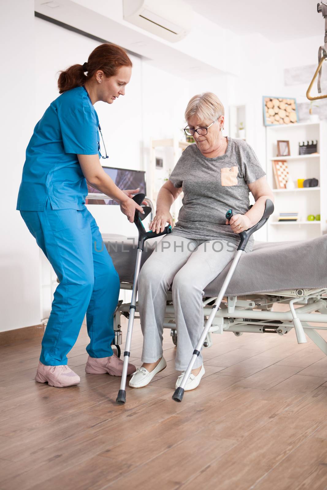 Nurse helping old woman to sit on bed in nursing home by DCStudio