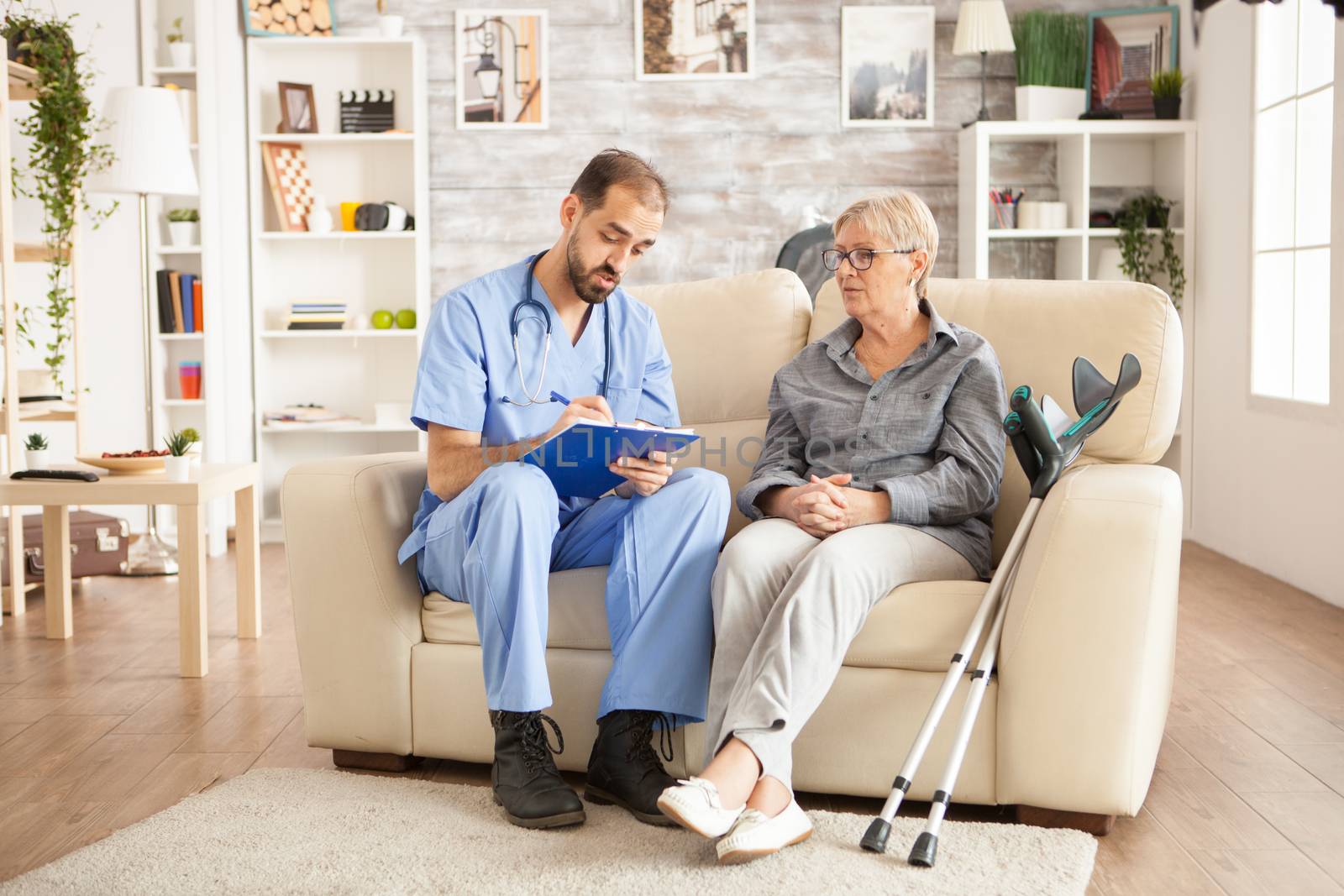 Male caregiver taking notes on clipboard while talking with senior woman in nursing home.