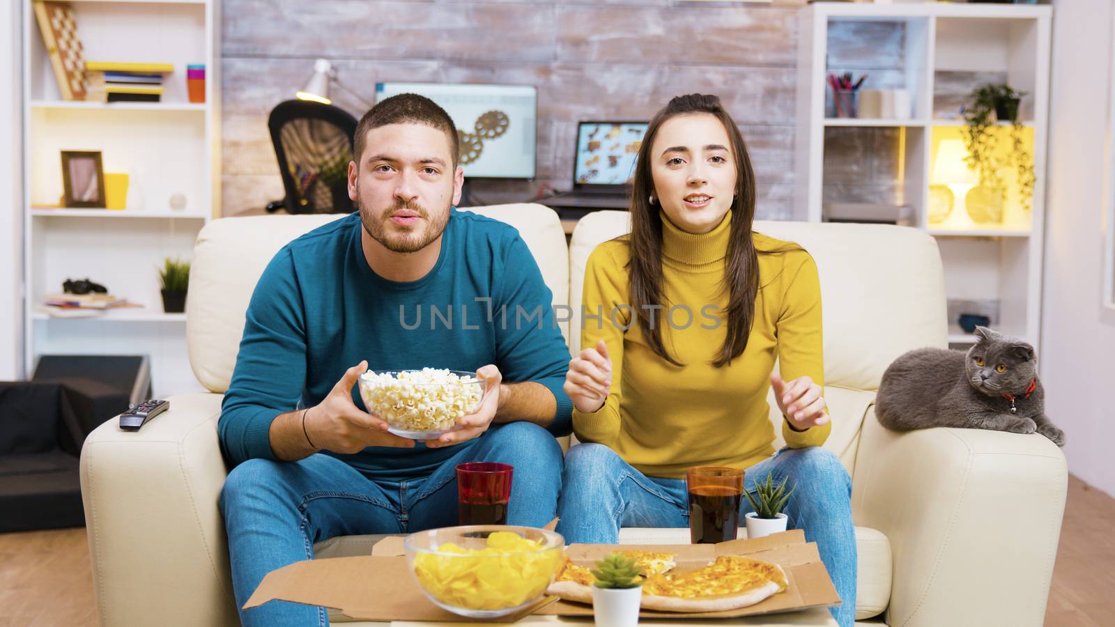 Excited couple sitting on the couch eating junk food and cheering up for their sport team while watching tv. Cat sitting on the sofa.