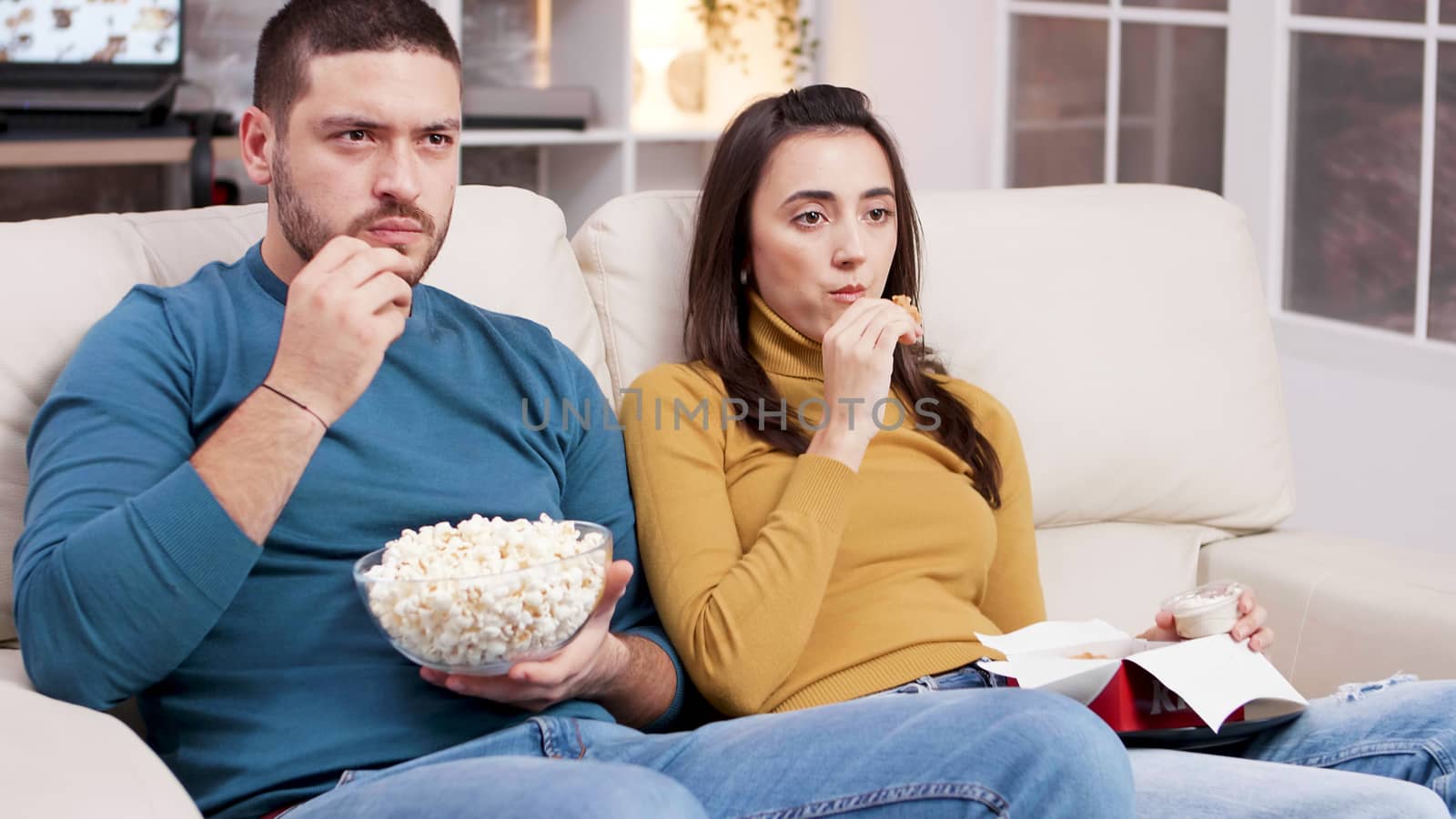 Couple sitting on couch eating fried chicken and popcorn while watching tv. Scared couple after a scary moment in the movie.