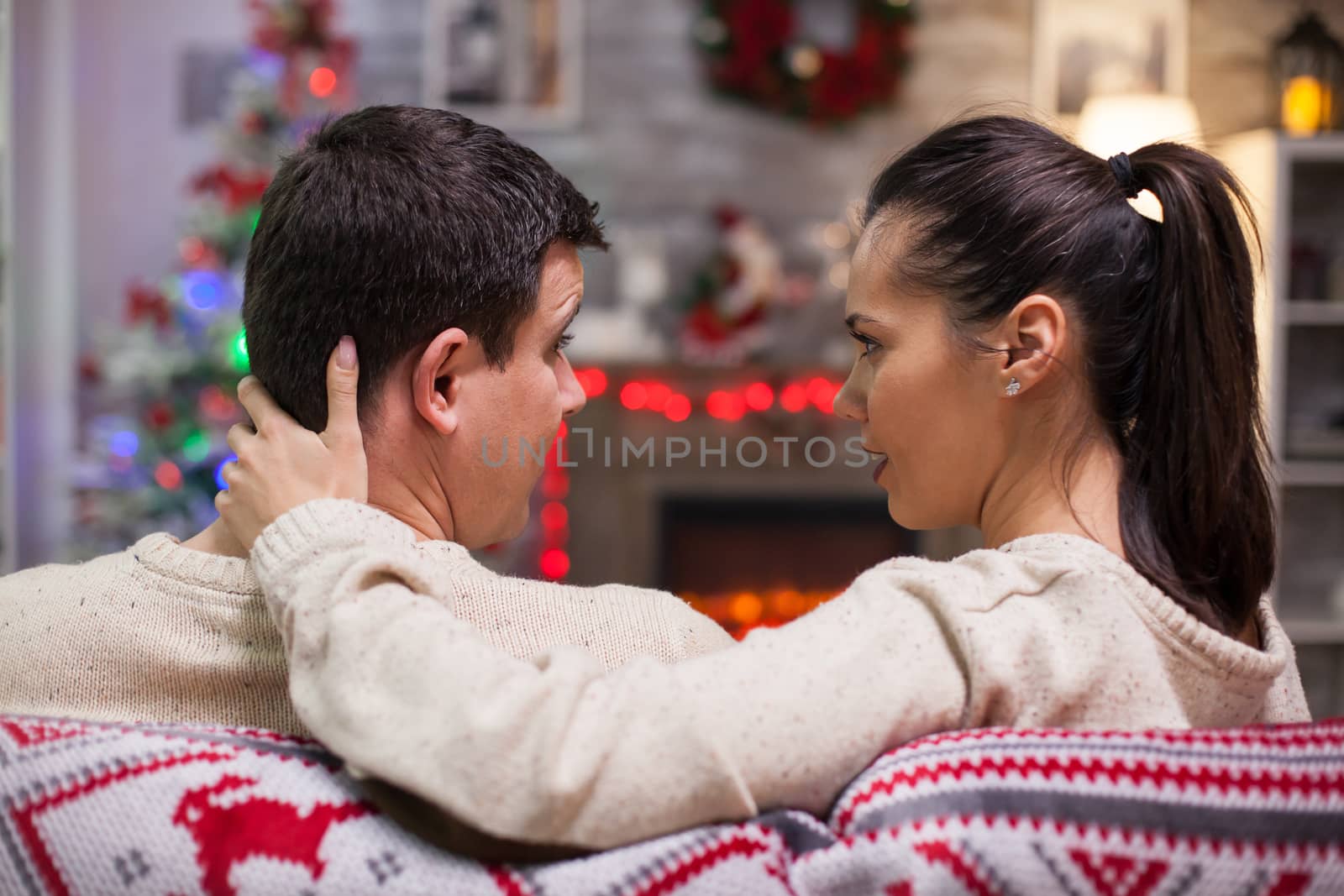 Back view of caucasian couple sitting on couch looking at each other in front of their fireplace.