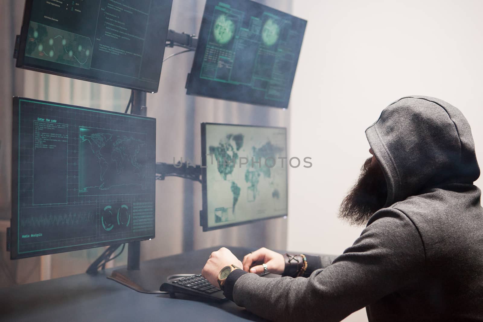 Side view of bearded hacker with a hoodie using multiple screens for cyberterrorism.