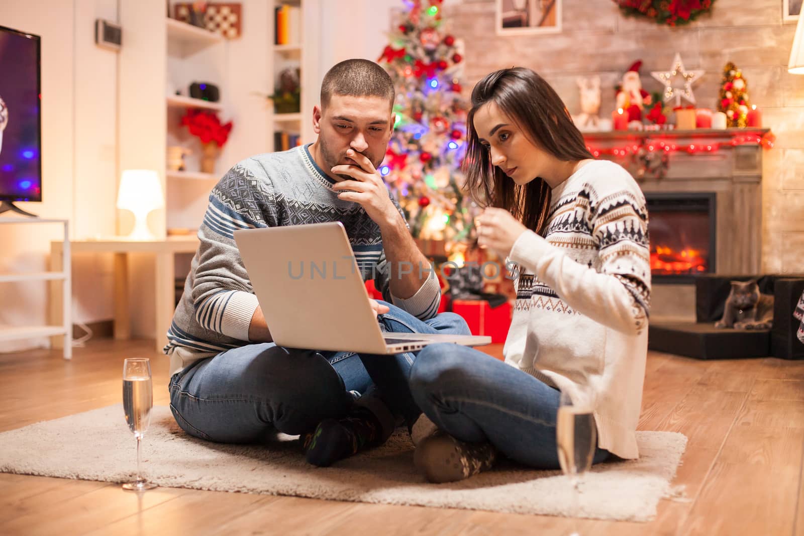 Happy couple looking for christmas gifts on internet using their laptop in front of fireplace.