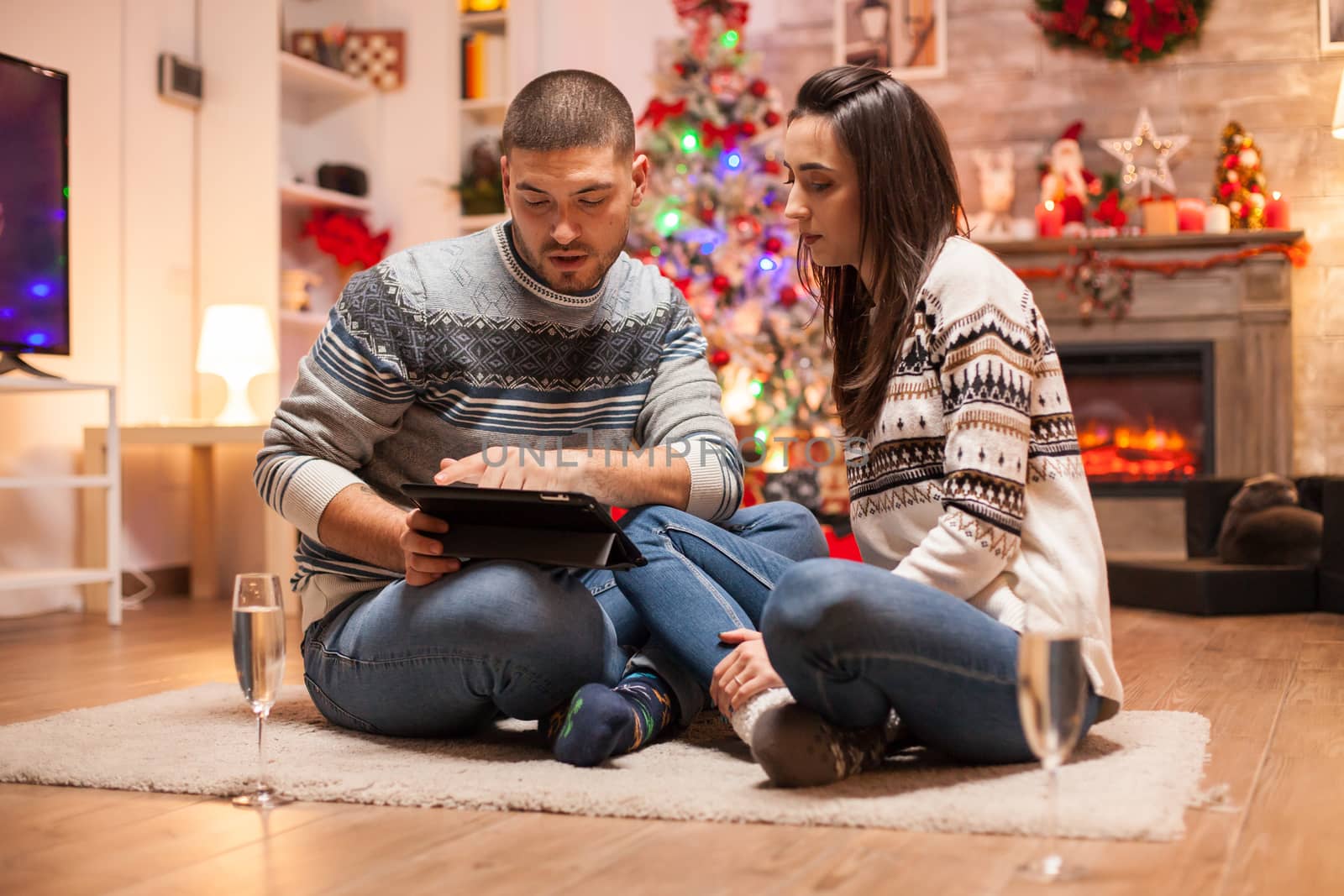Couple in love sitting in front of christmas tree looking on tablet computer.