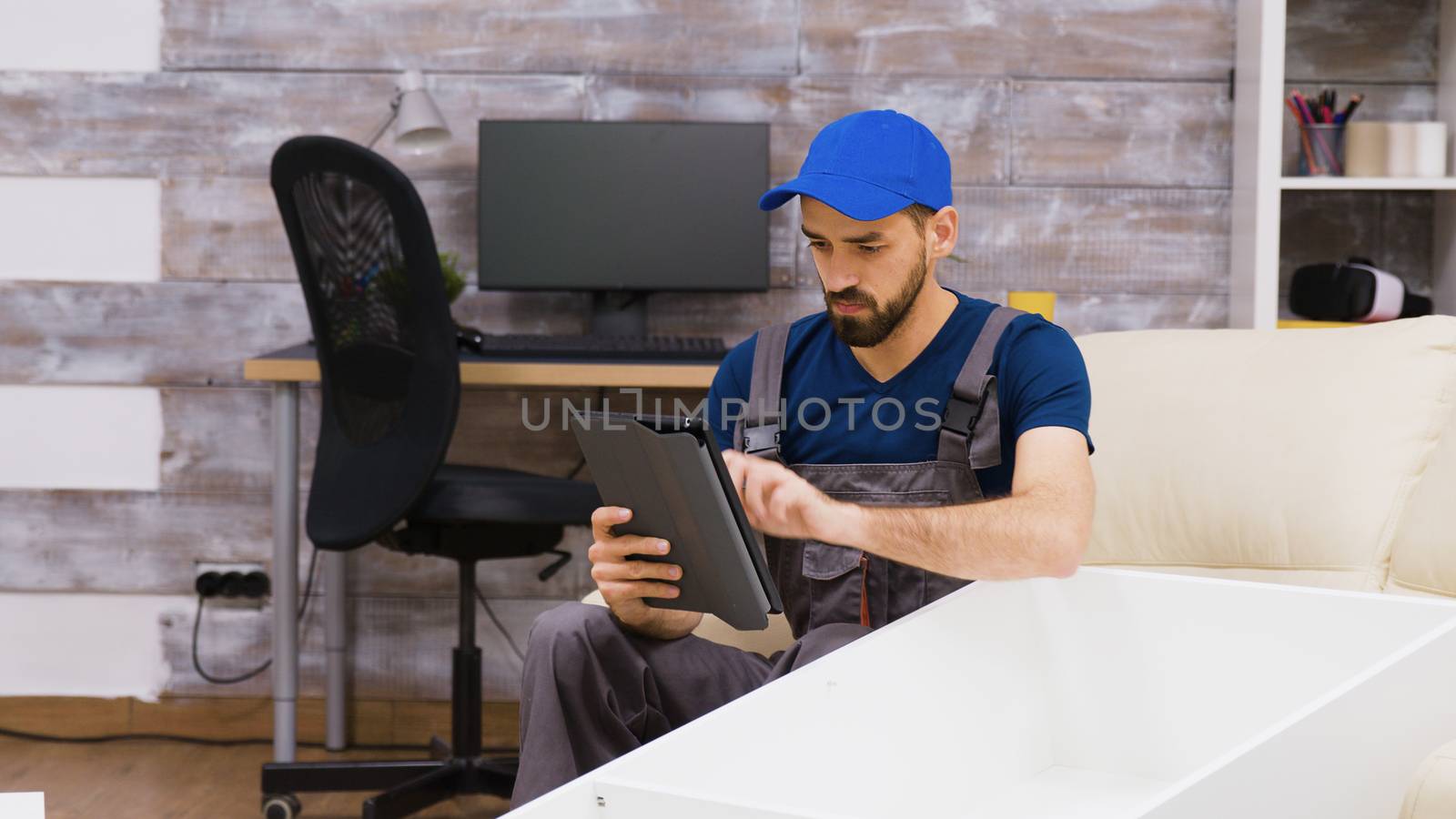 Male worker in overalls wearing a cap reading furniture assembly from a tablet computer.