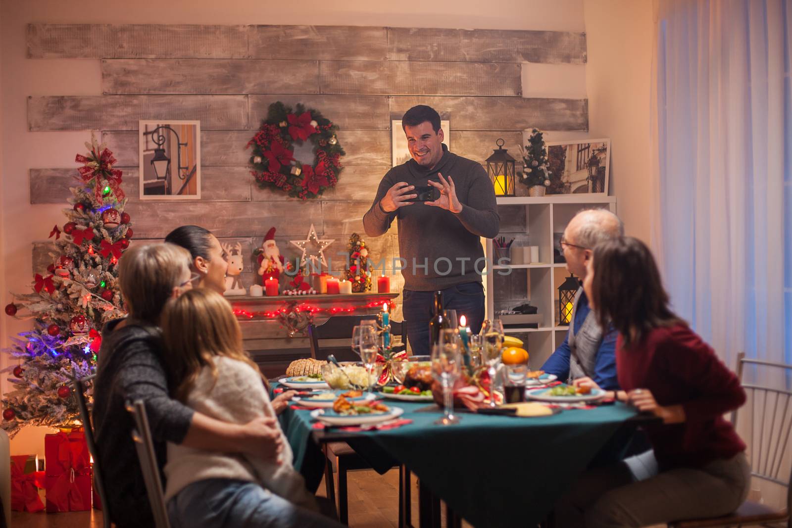 Cheerful man using smartphone to take photos of his family at christmas celebration.