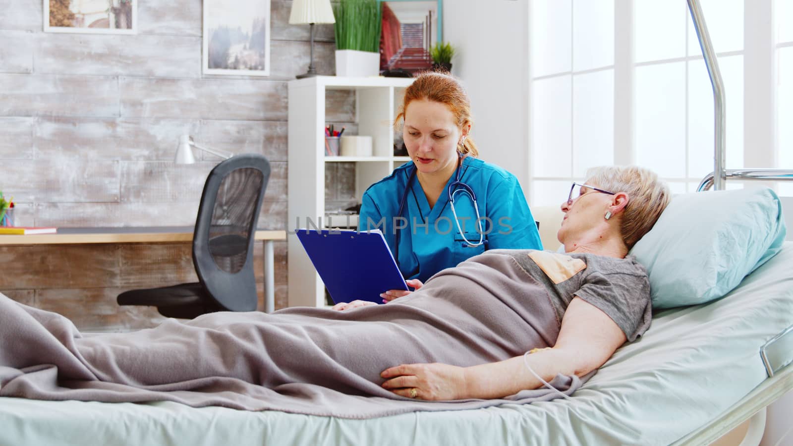 Social worker consulting an elderly disabled woman who lies in hospital bed. The caregiver uses a clipboard to take pensioners notes