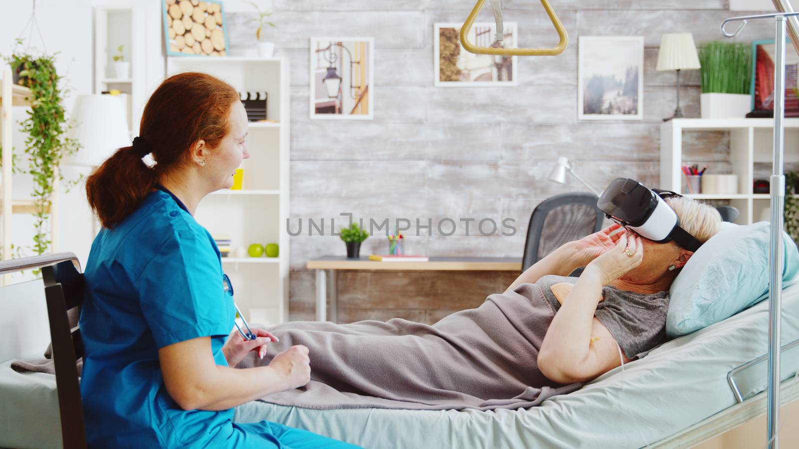 Female nurse in retirement home giving an old disabled lady a VR headset to ease the pain and distract her while she is lying in hospital bed
