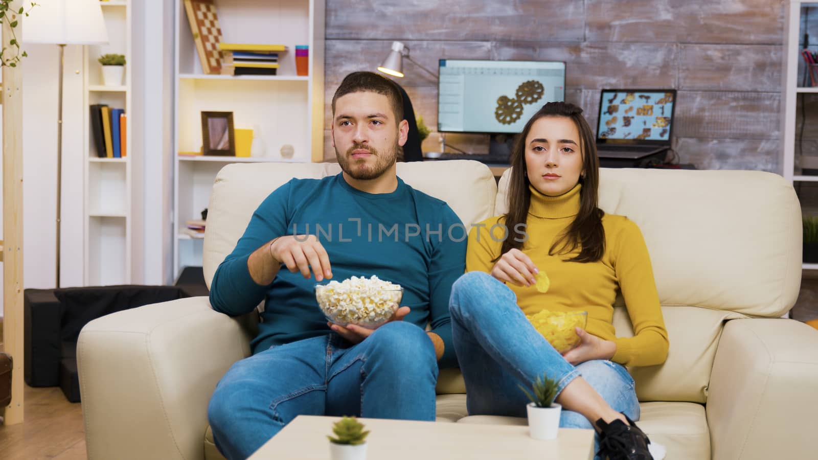 Beautiful young girl laying on her boyfriend's shoulder while watching tv and eating chips. Boyfriend eating popcorn.