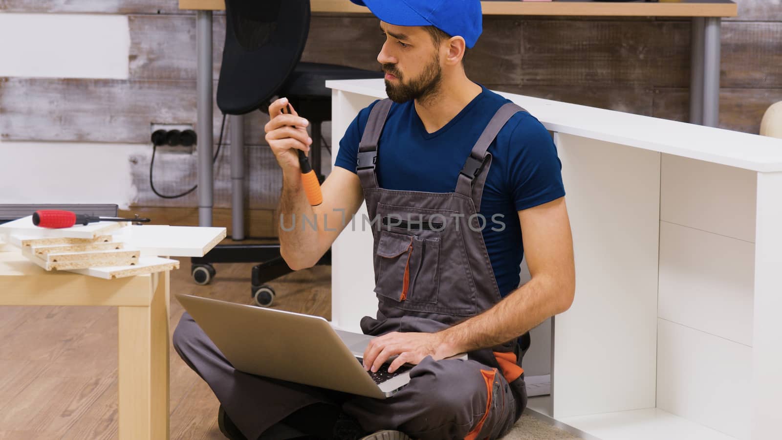 Furniture assembly worker wearing overalls searching instructions using his laptop. Worker holding screwdriver.