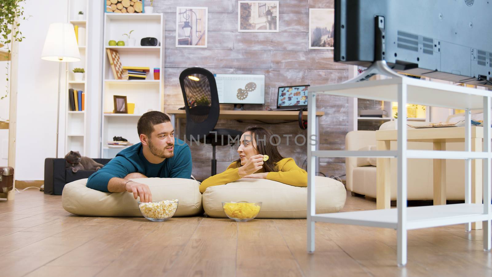 Couple talking with each other watching tv sitting on the pillows for the floor while their cat is relaxing in the background.