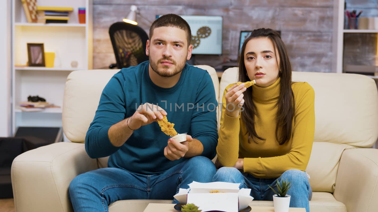 Young couple sitting on couch eating fried chicken while watching tv.