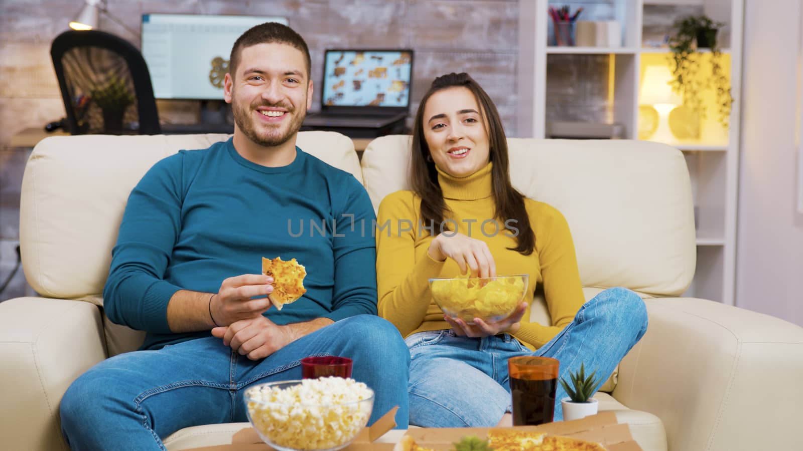 Cheerful bearded man laughing while watching a movie with his girlfriend and eating pizza. Popcorn and soda on coffee table.