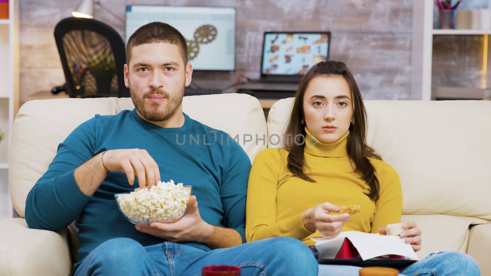 Scared couple watching tv eating pizza and popcorn sitting on couch. Couple eating junk food.