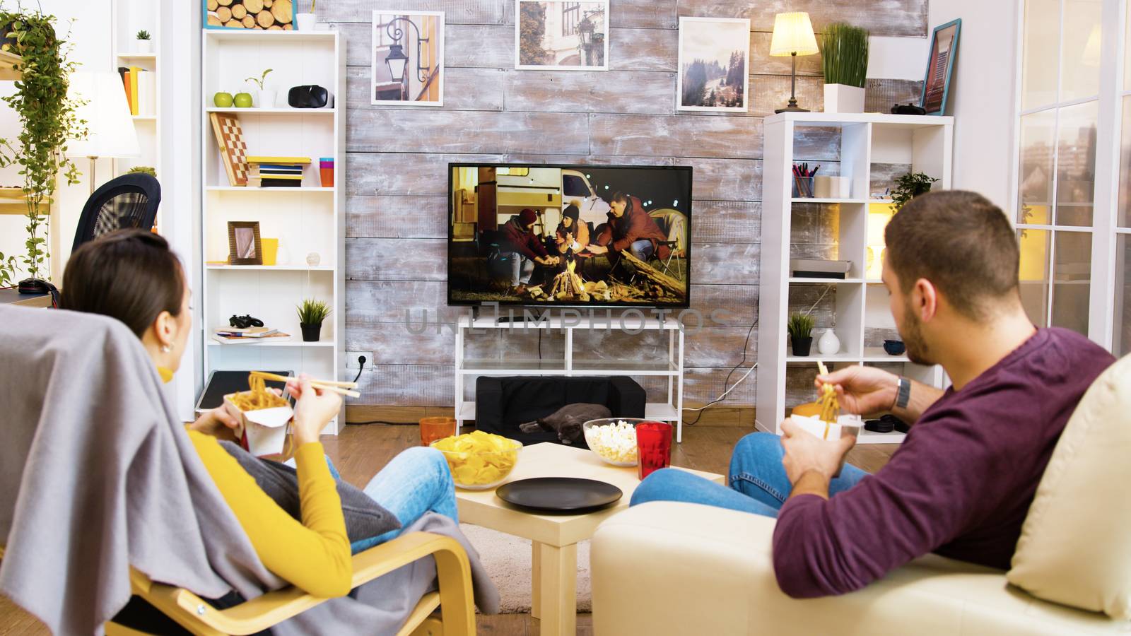 Back view of bearded man with his girlfriend sitting on chairs eating noodles with chopsticks in front of tv. Cat sleeping.