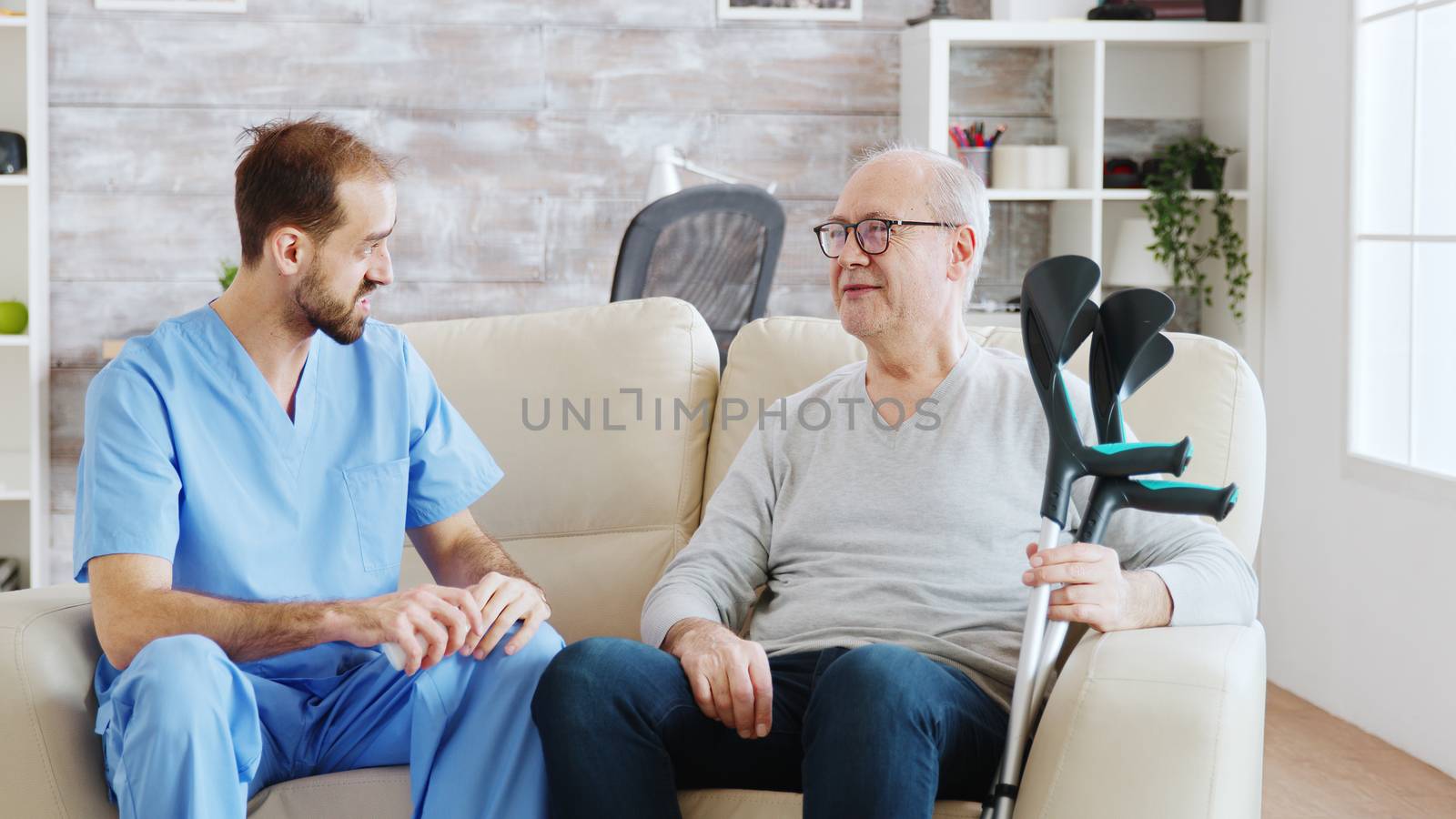 In retirement home male nurse is talking with an old disabled man with crutches next to him. The caregiver is consulting pensioner about drugs dosage