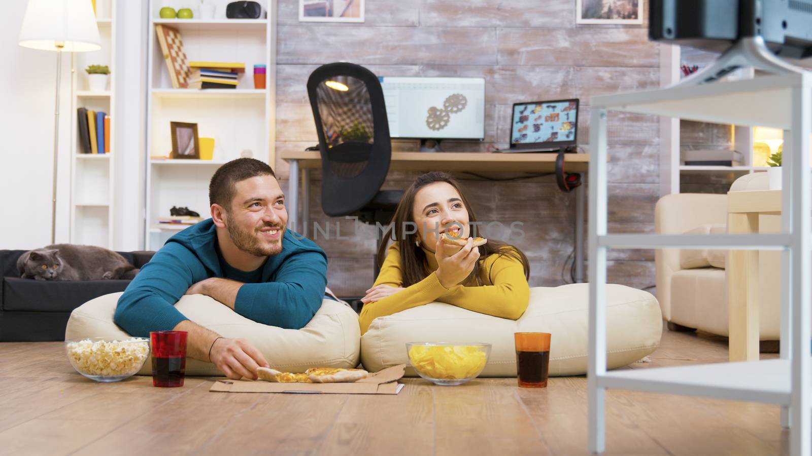 Happy caucasian couple eating pizza sitting on pillows for the floor watching tv while their cat is relaxing in the background.