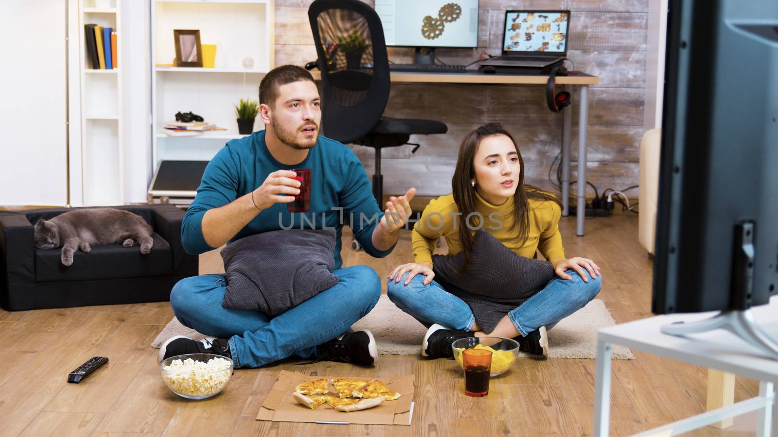 Young couple sitting on the floor cheering up, eating junk food while watching sport championship on tv and cat sleeping.