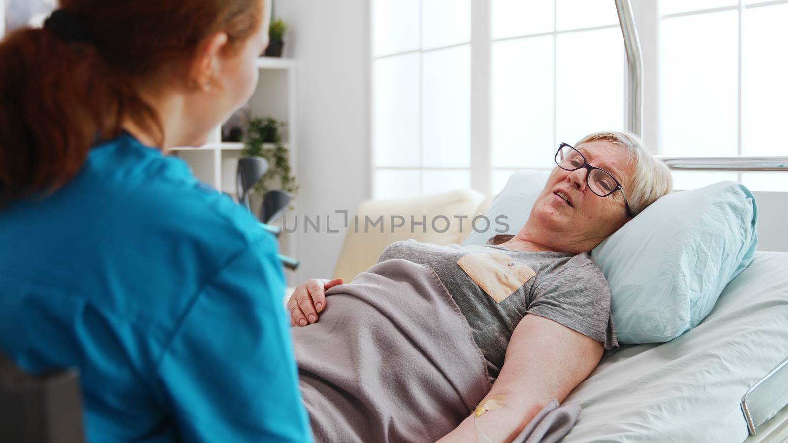 In nursing home old sick lady lying in hospital bed talks with a female nurse. Over the shoulder shot