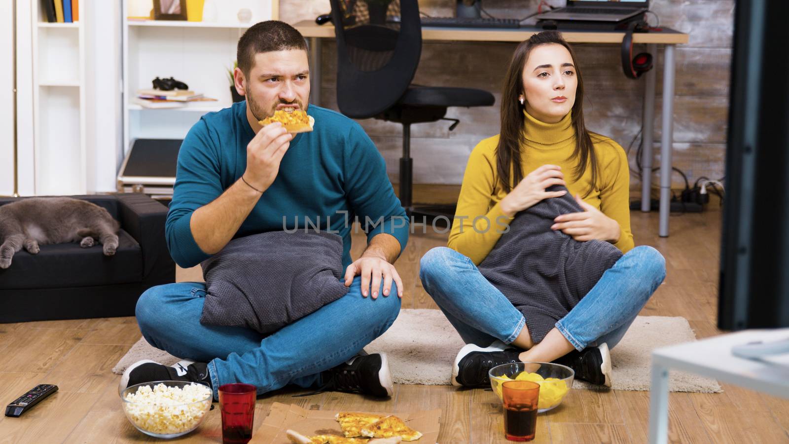 Disappointed couple sitting on the floor while watching sport on tv and eating pizza with their cat sleeping.