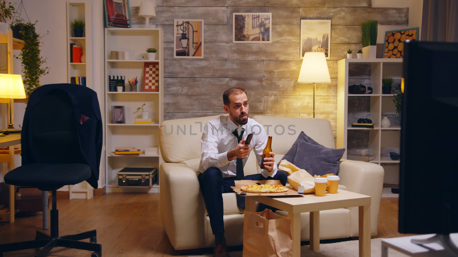 Businessman with tie eating pizza and watching tv after work. Drinking beer.