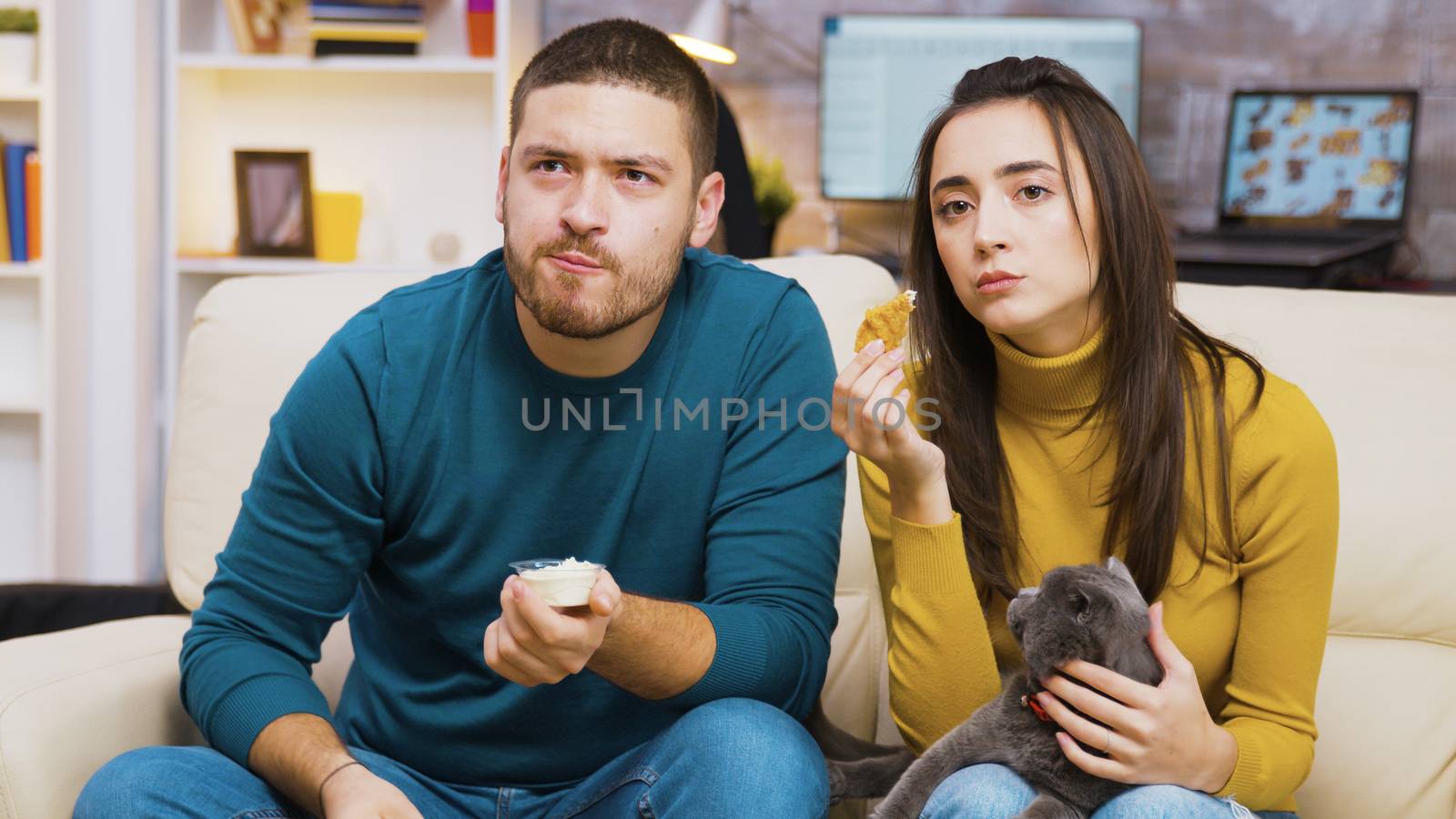 Concentrated young couple sitting on couch eating fried chicken while watching tv with the cat on girl lap.