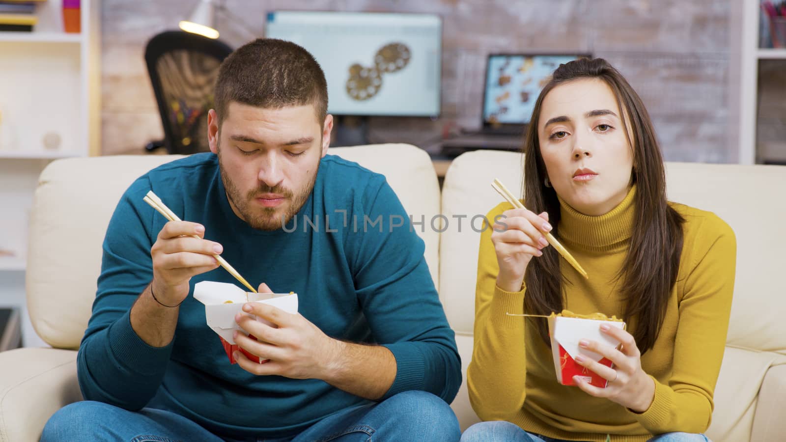 Bearded man and his girlfriend eating noodles with chopsticks while watching tv.