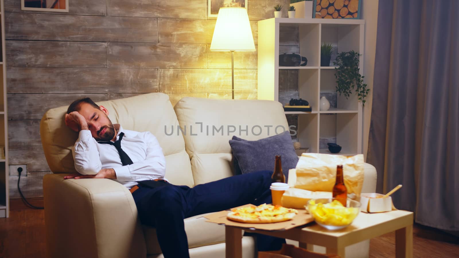 Overworked businessman sleeping on the couch with tv turned on and junk food on the table.