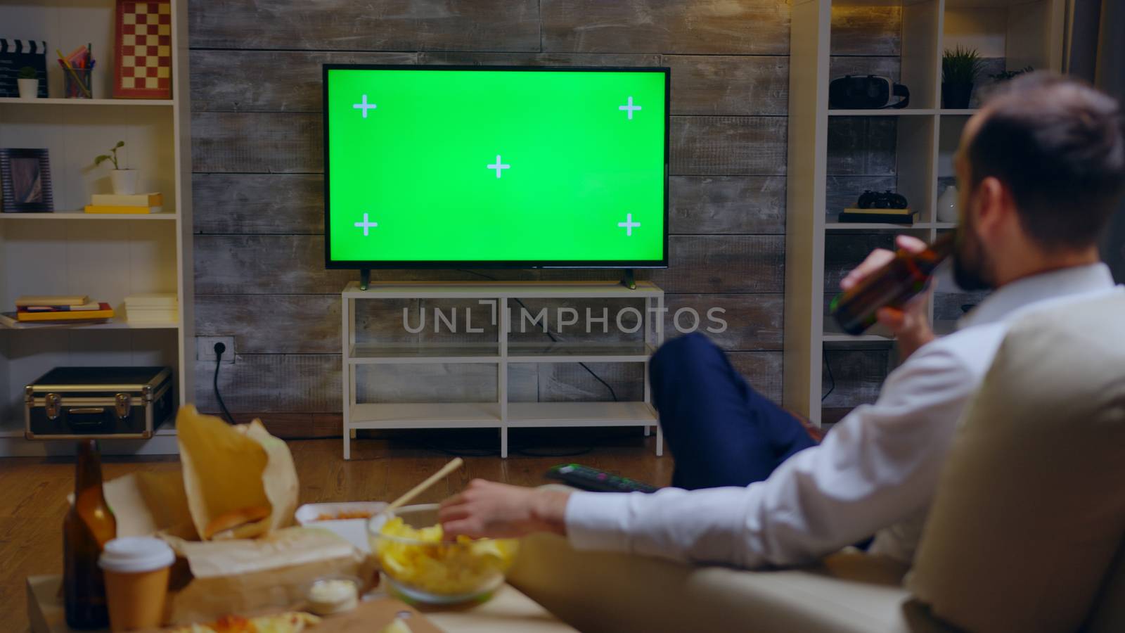 Back view of young entrepreneur relaxing watching tv. Green screen and junk food.