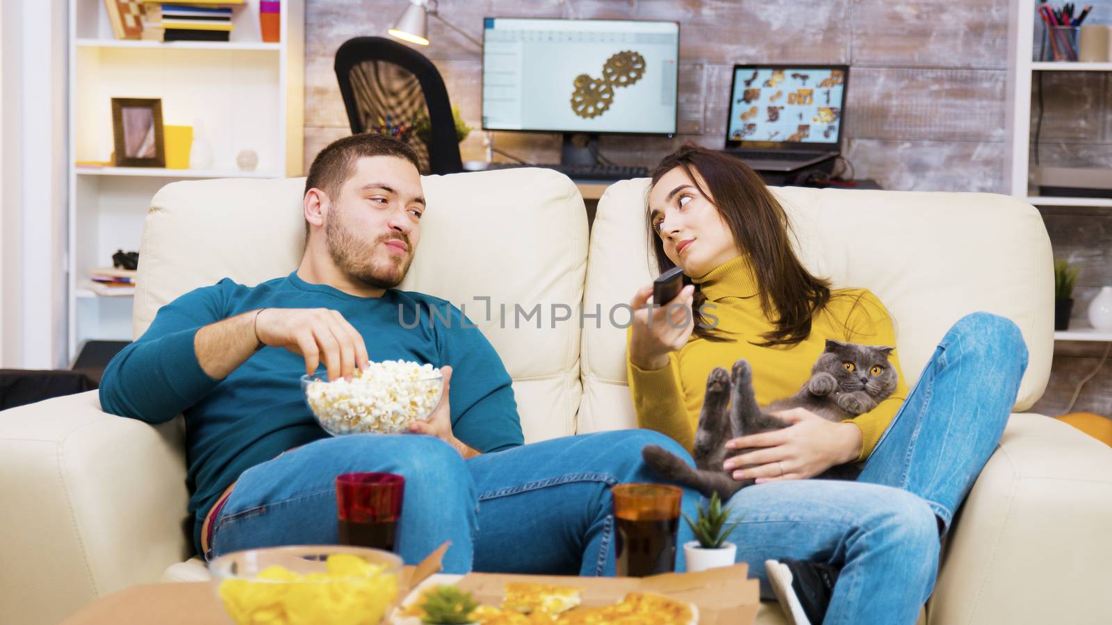 Bored girl sitting on the couch with the cat in her lap and boyfriend next to her is using tv remote control.