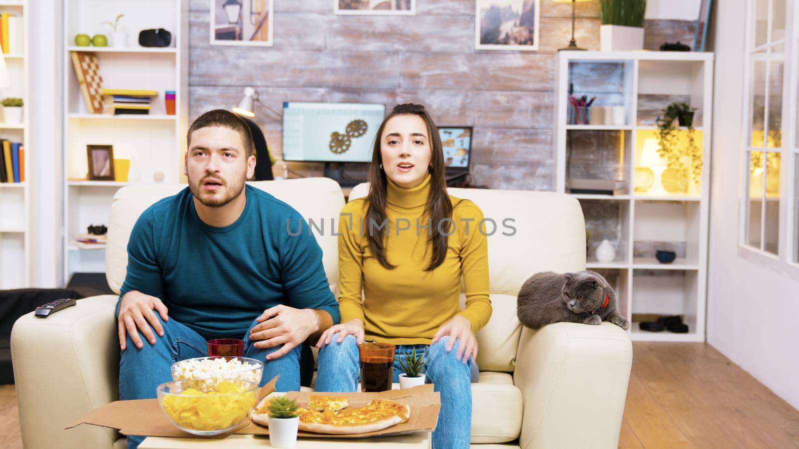 Disappointed couple after their favorite football team lost the game. Cat laying on sofa. Popcorn, pizza and soda on coffee table.