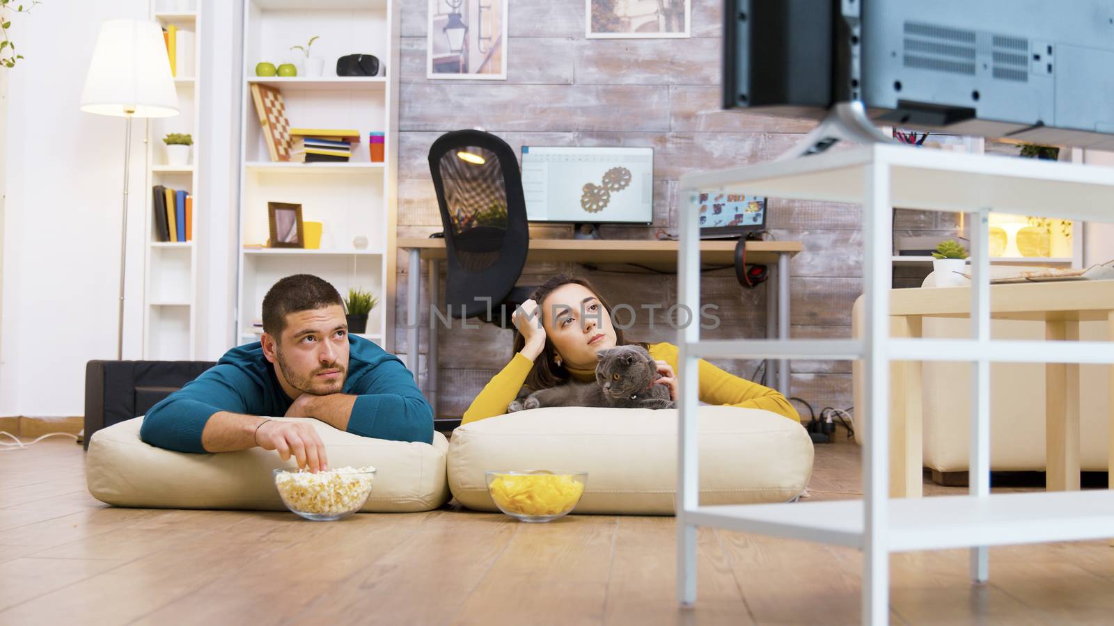 Young couple watching tv with their cat sitting on pillows for the floor while eating chips and popcorn.