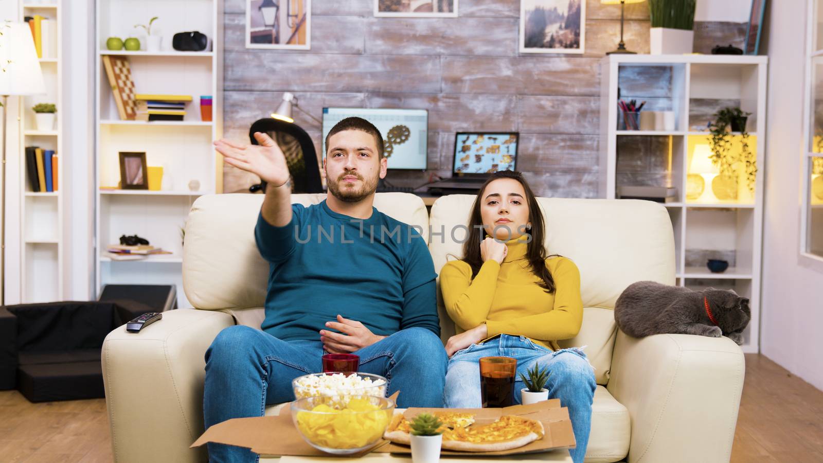 Bearded man changing tv channels with hand gestures while his girlfriend and the cat are next to him on the couch.