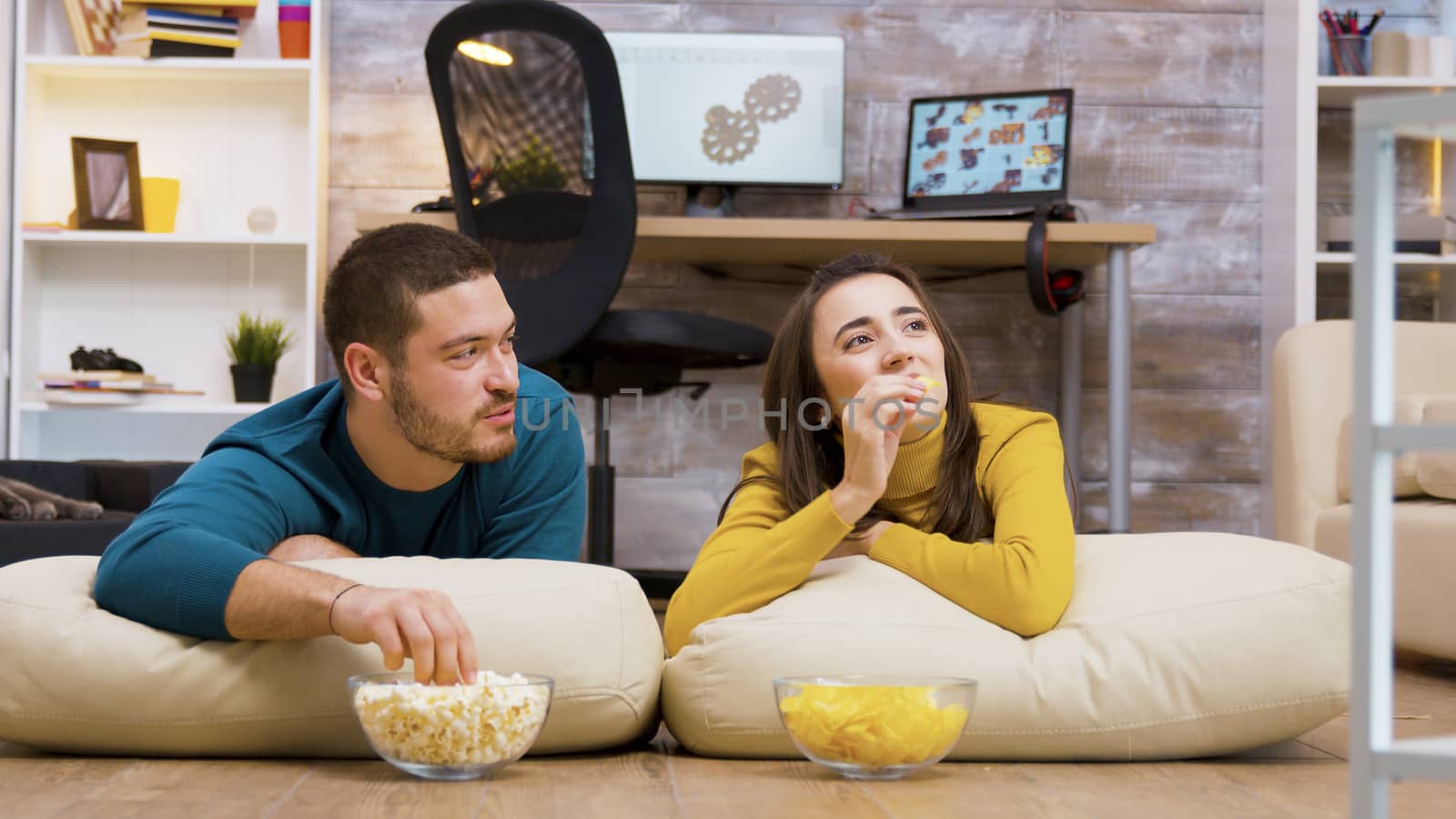 Cheerful couple watching tv sitting on pillows for the floor eating chips and popcorn with their cat in the background.