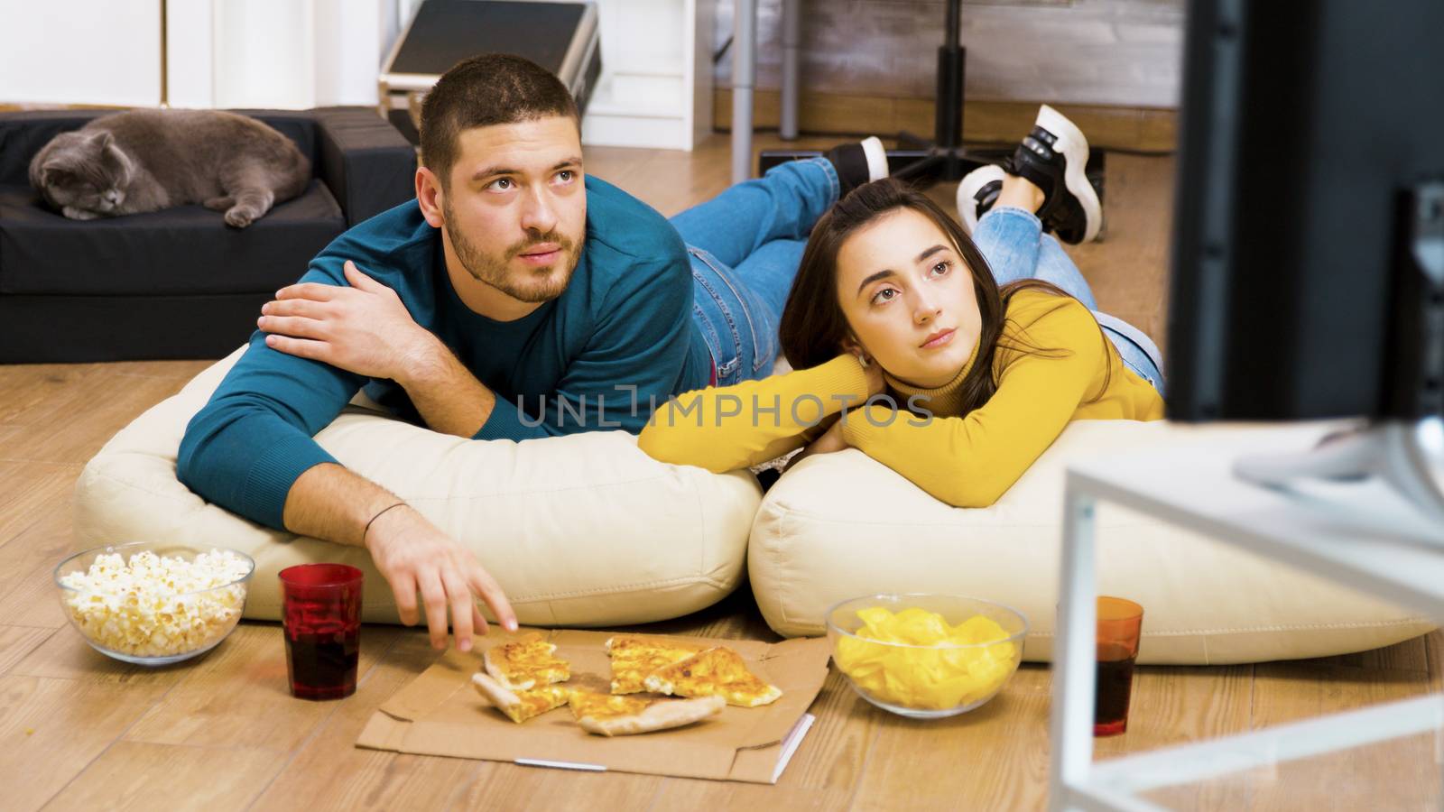 Attractive couple relaxing on pillows for the floor watching tv and eating pizza with their cat sleeping in the background.